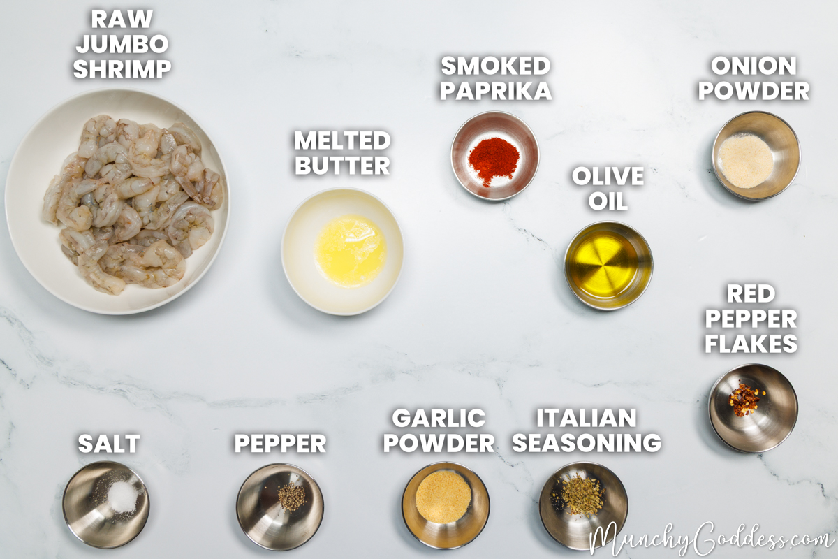 Air fryer shrimp recipe ingredients in various dishes on a white marble surface: raw jumbo shrimp, melted butter, smoked paprika, olive oil, onion powder, salt, ground black pepper, garlic powder, Italian seasoning, and red pepper flakes.