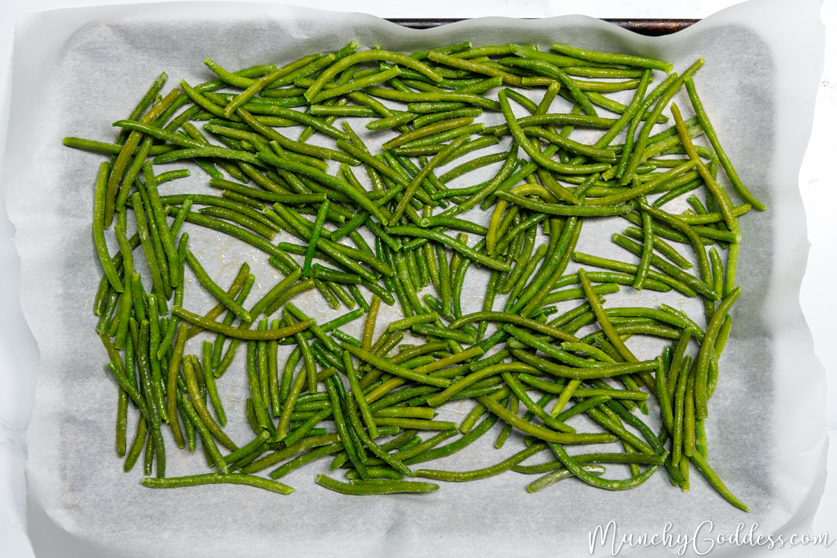 Frozen green beans coated with olive oil spread out on a sheet pan lined with parchment paper.