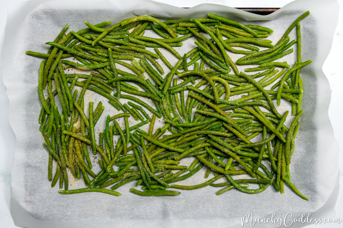 Salt, ground black pepper, garlic powder, and onion powder added on top of frozen green beans coated in olive oil on a sheet pan lined with parchment paper.