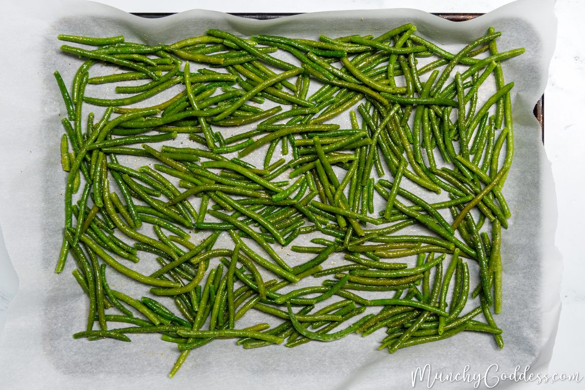 Frozen green beans evenly spread out and coated in olive oil, salt, ground black pepper, garlic powder, and onion powder on a sheet pan lined with parchment paper.