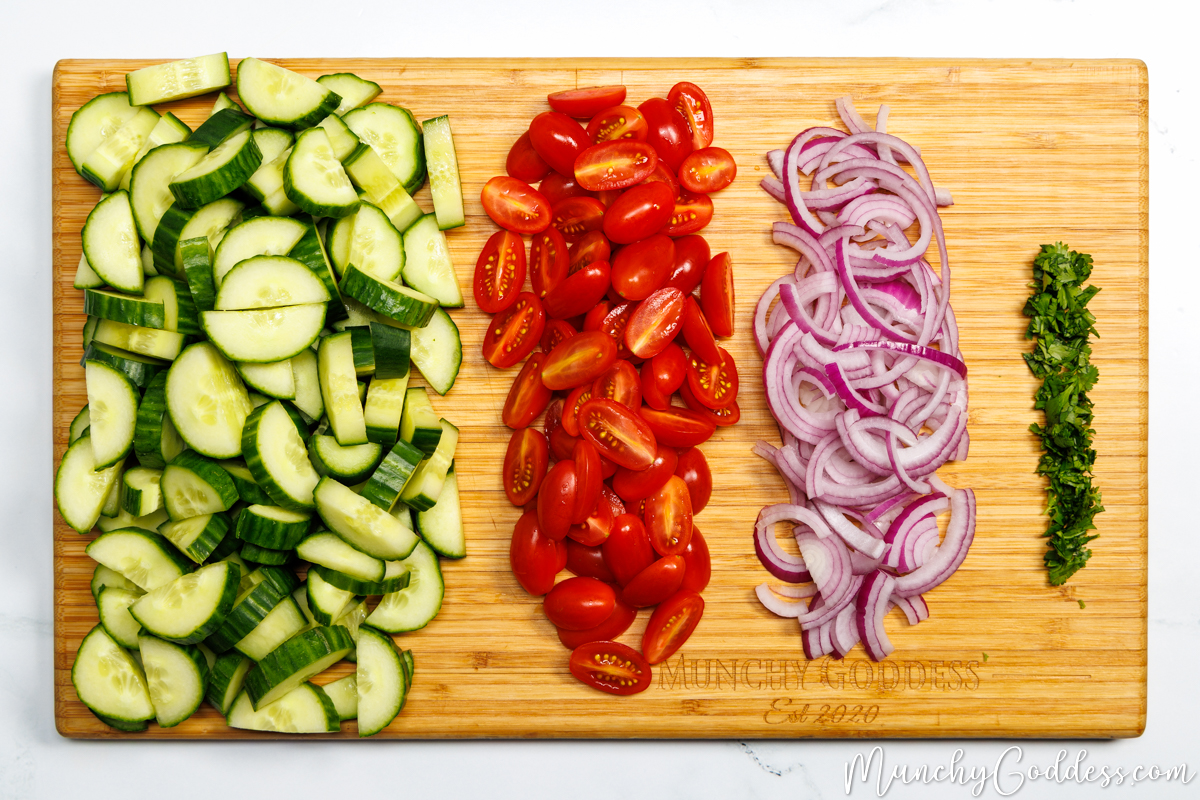 Sliced cucumbers, halved tomatoes, thinly sliced red onion, and finely chopped parsley in neat columns on a wood cutting board.