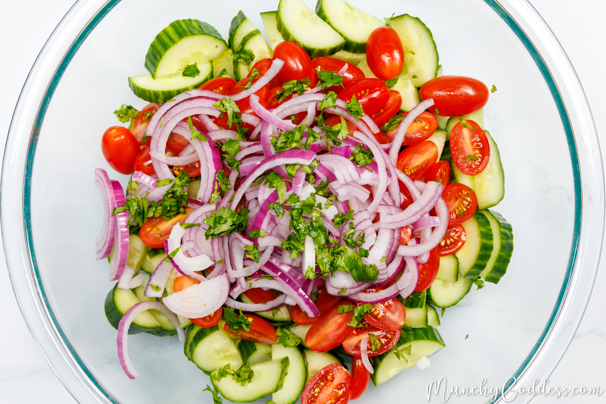 Sliced cucumbers, halved tomatoes, thinly sliced red onion, and finely chopped parsley in a large glass bowl.