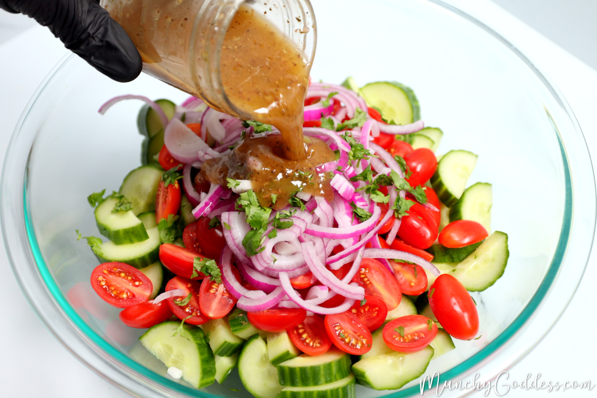 Homemade salad dressing being poured over cucumbers, cherry tomatoes, red onions and fresh parsley in a large glass bowl.