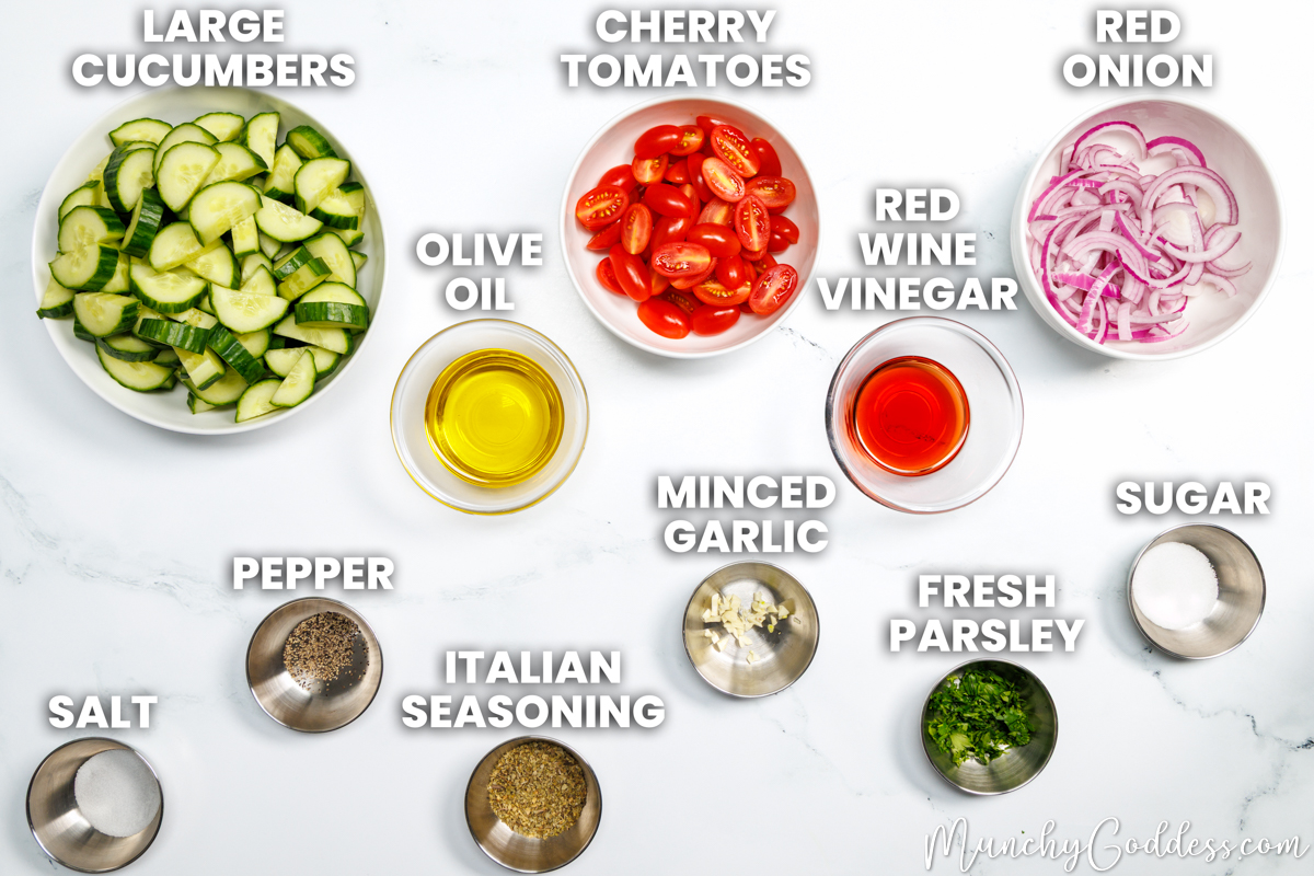 Cucumber tomato salad recipe ingredients in various dishes on a white marble surface: large cucumbers, olive oil, cherry tomatoes, red wine vinegar, red onion, salt, ground black pepper, Italian seasoning, minced garlic, fresh parsley, and granulated sugar.