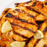 Grilled chicken thighs with dark, parallel sear marks garnished with lemon slices on an off-white plate.