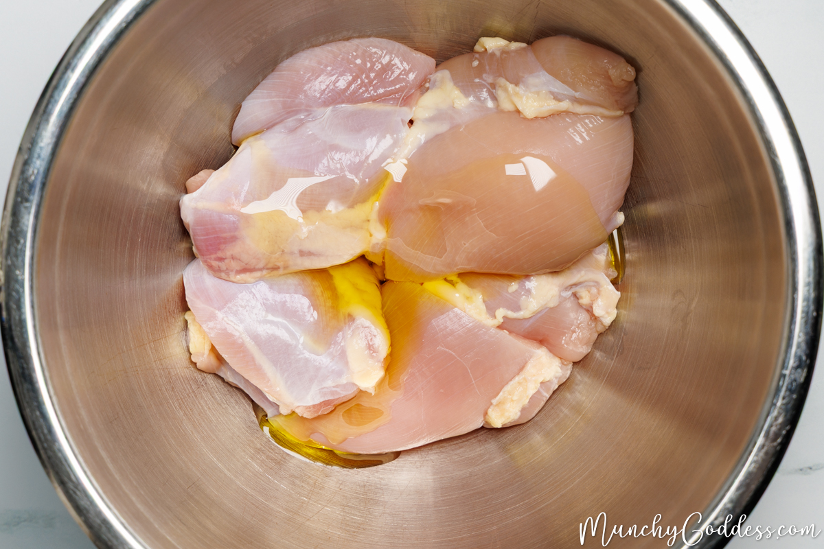 Boneless skinless chicken thighs drizzled with olive oil in a silver mixing bowl.