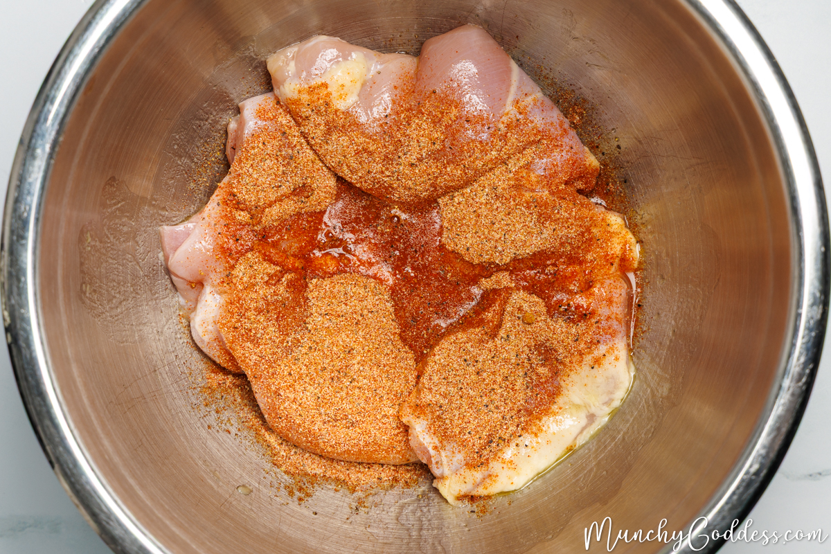 Boneless skinless chicken thighs coated with olive oil and topped with seasoning mix and fresh lemon juice in a silver mixing bowl.