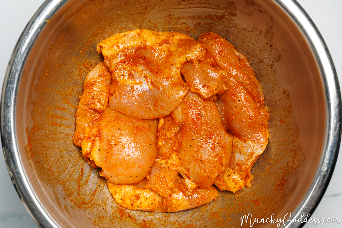 Boneless skinless chicken thighs tossed and evenly coated in an olive oil, seasoning, and fresh lemon juice mixture in a silver mixing bowl.