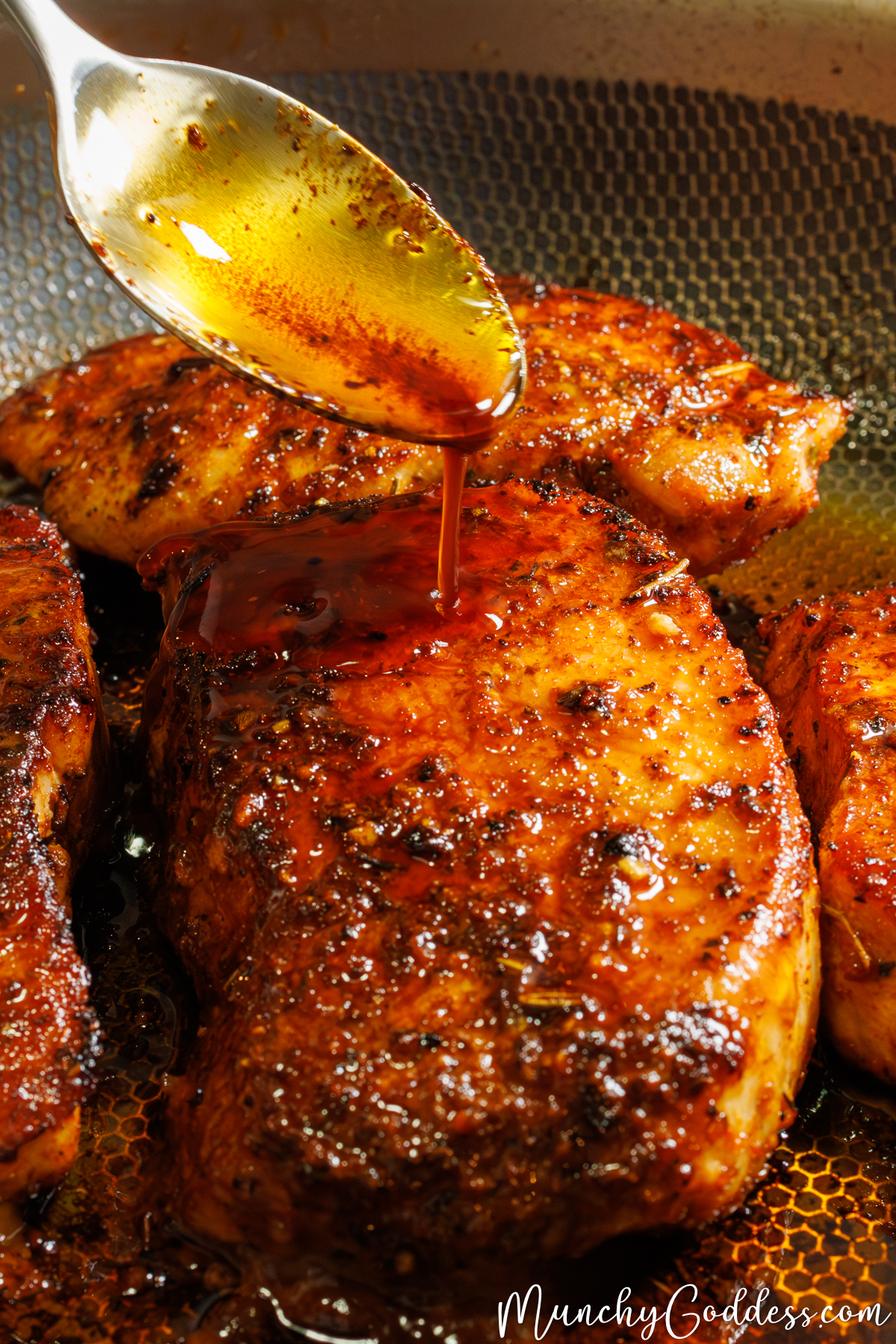 A matte silver spoon pouring a shimmering pan butter sauce over a golden brown lemon pepper pork chop in a textured skillet.