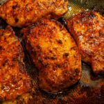 Golden brown pan-seared lemon pepper pork chops with a glistening glaze in a textured skillet with savory drippings.