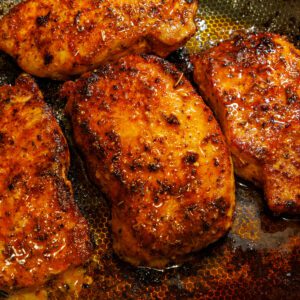 Golden brown pan-seared lemon pepper pork chops with a glistening glaze in a textured skillet with savory drippings.