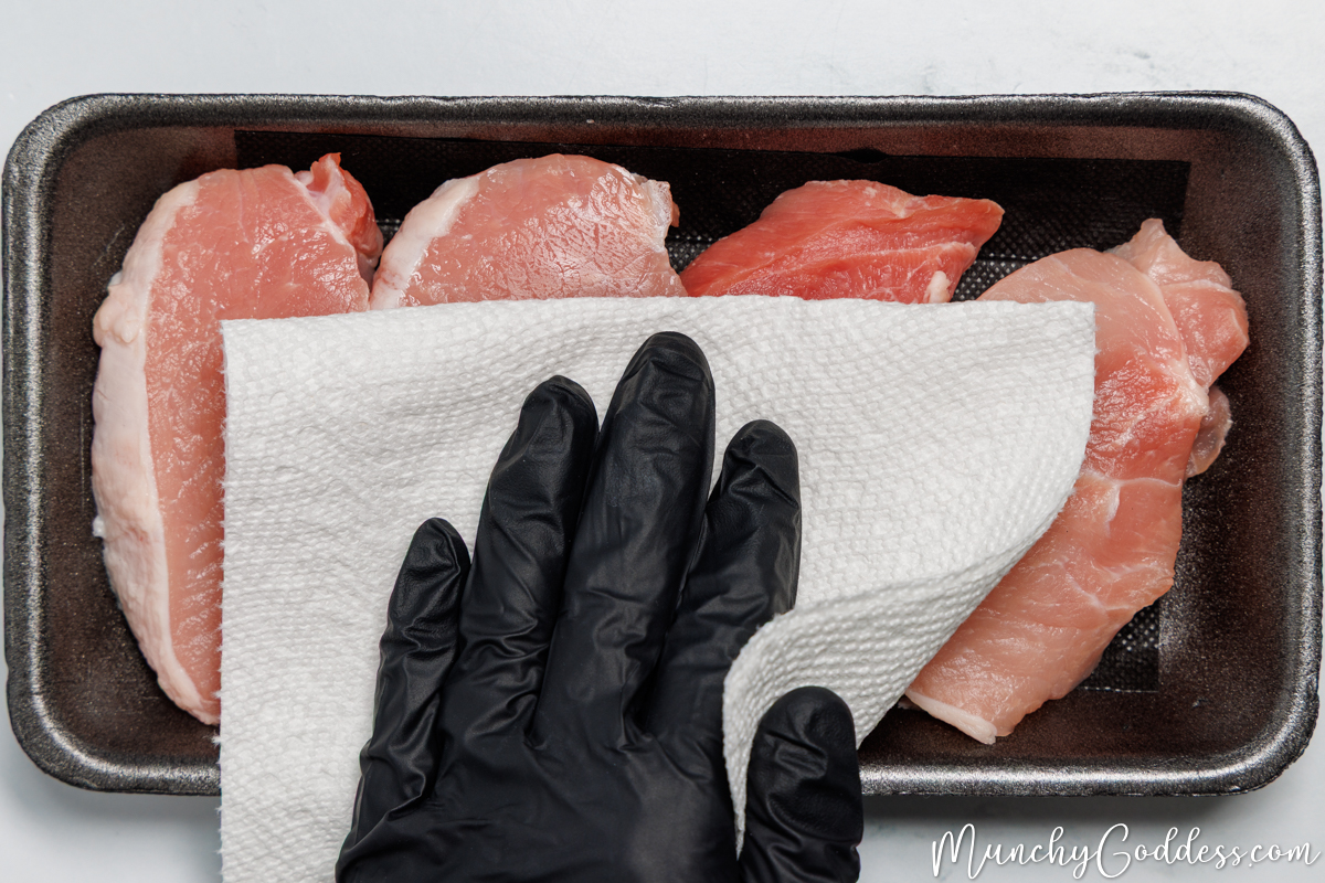 Boneless pork chops in a black foam meat tray being patted dry with a paper towel.