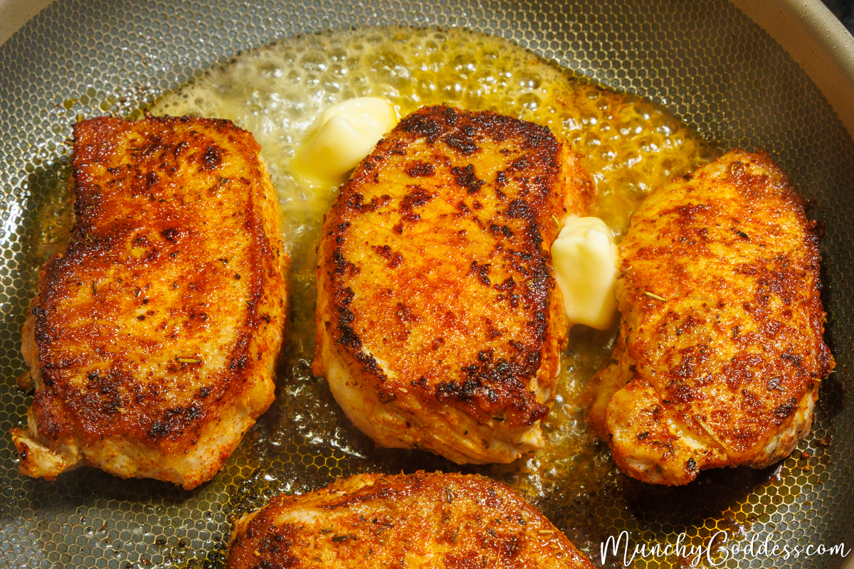 Golden brown, spice-rubbed boneless pork chops searing in a textured skillet, surrounded by melting pats of butter and hot bubbling oil.