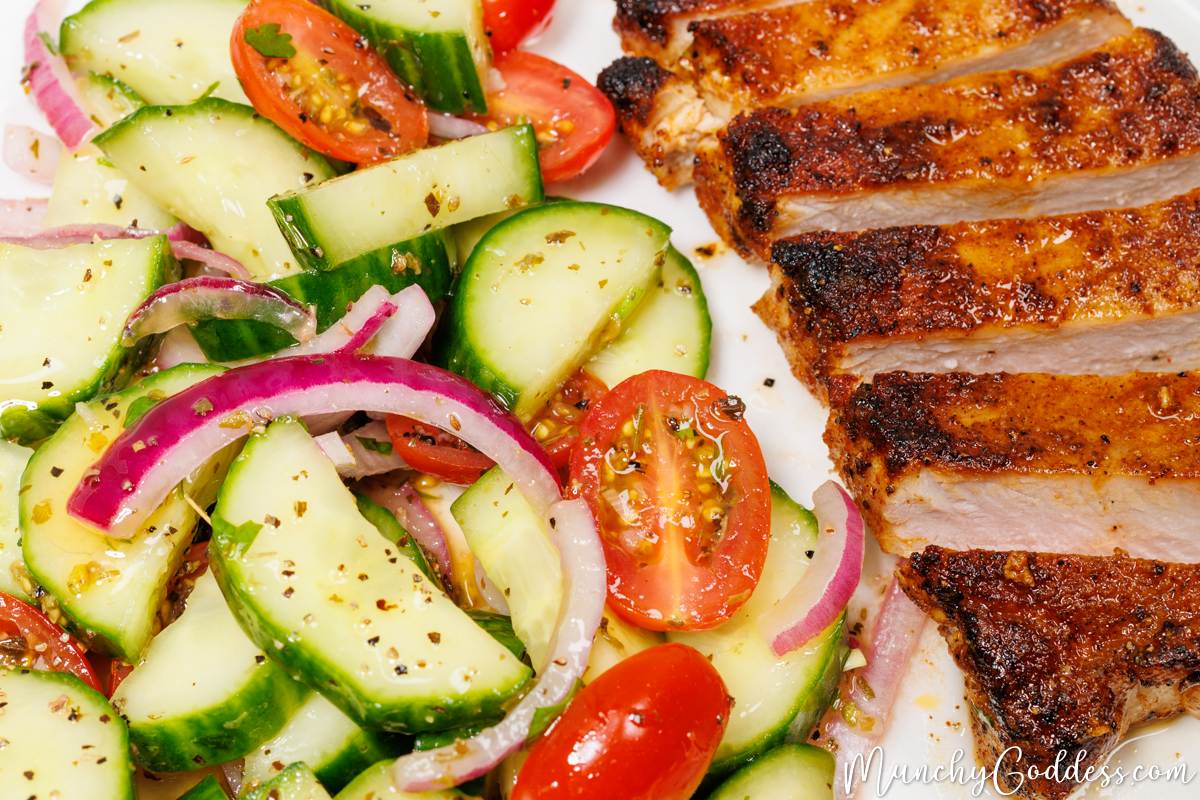 Cucumber tomato salad with dressing next to a juicy sliced boneless pork chop on a white plate.