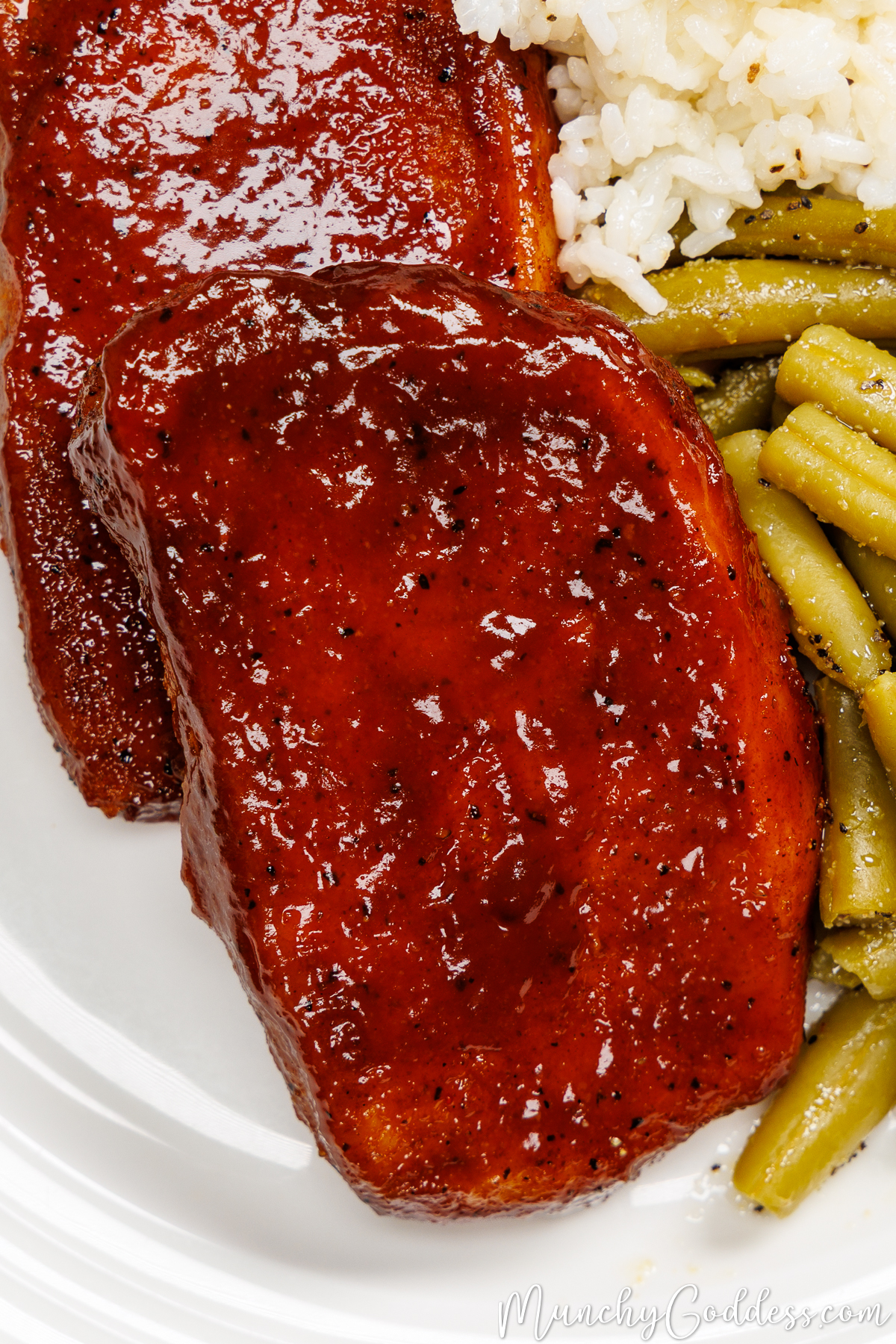 Glistening baked bbq pork chops with green beans and rice on an off-white plate.