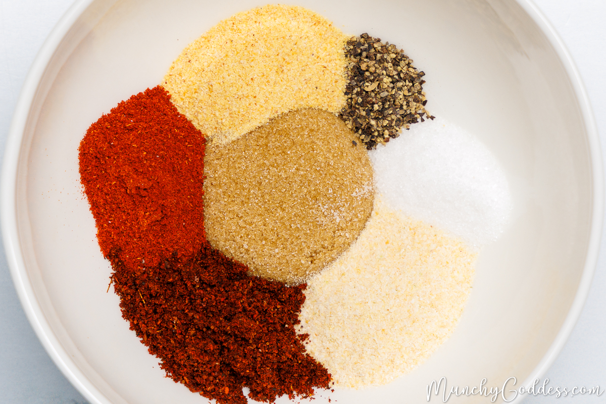 Salt, ground black pepper, onion powder, garlic powder, smoked paprika, chili powder, and brown sugar neatly arranged in an off-white bowl.