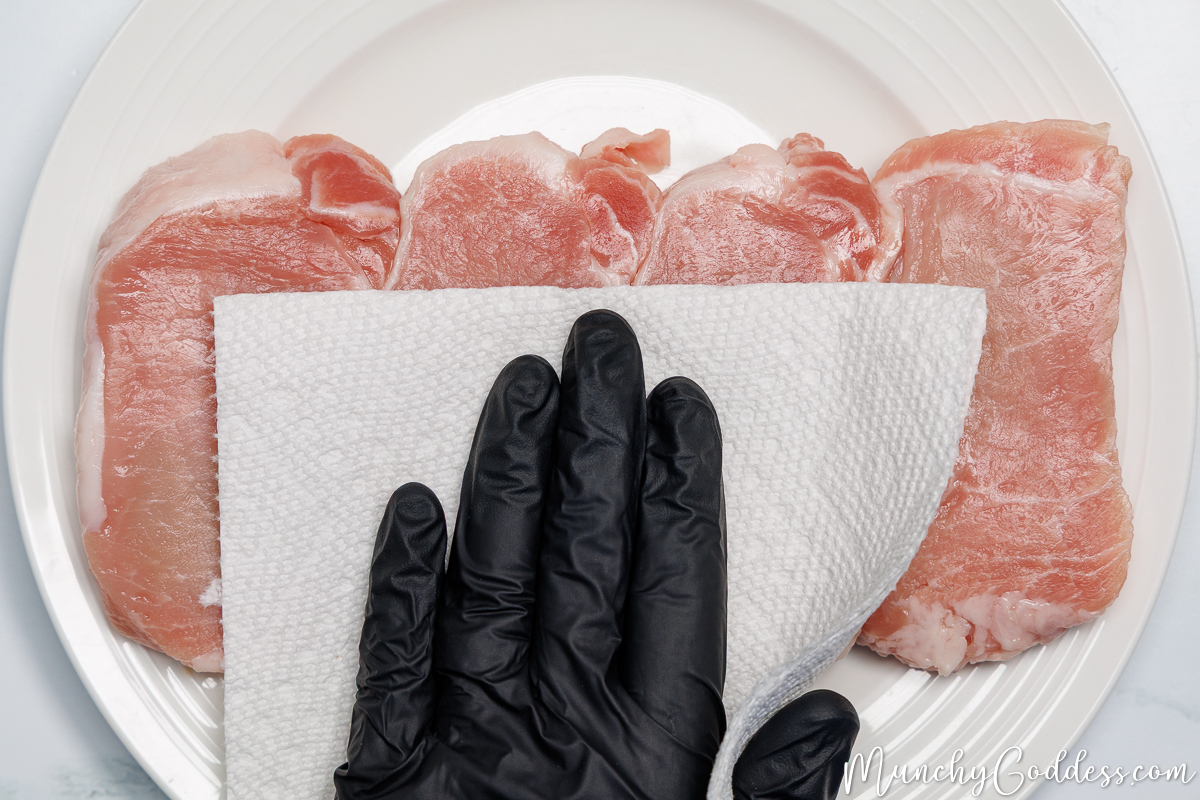 Boneless pork chops being patted dry with a paper towel on an off-white plate.