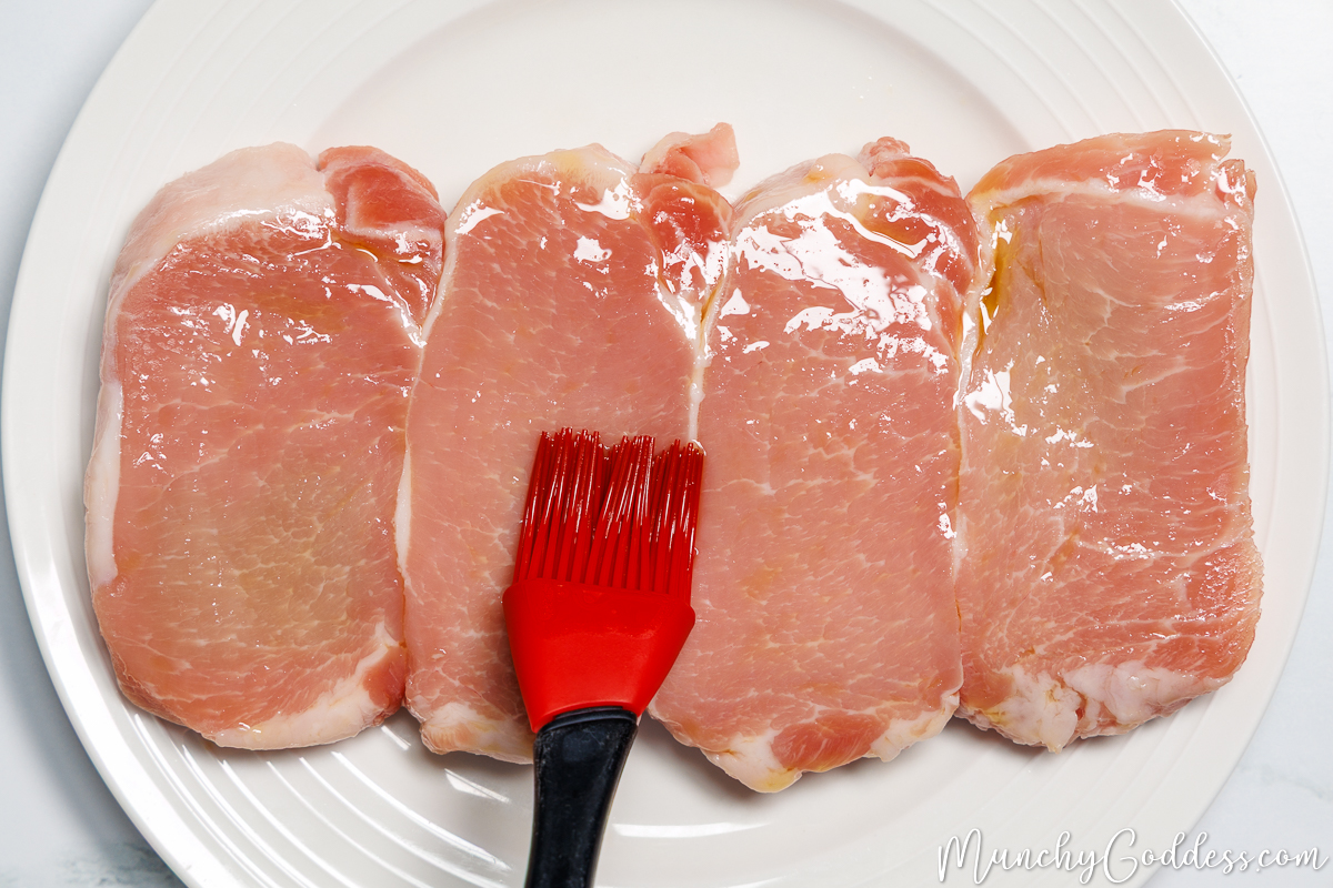 Boneless pork chops being brushed with olive oil with a red and black basting brush on an off-white plate.