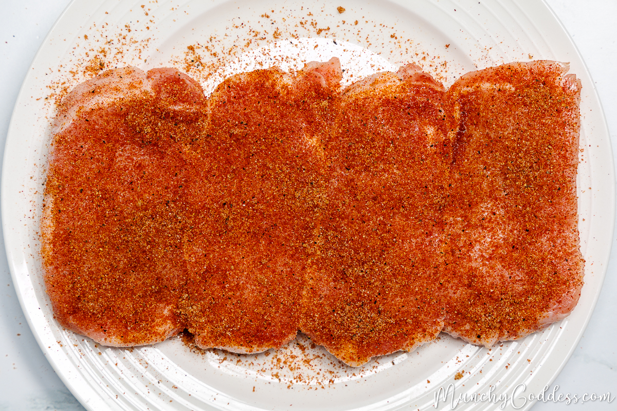 Seasoning sprinkled on top of oil-brushed boneless pork chops on an off-white plate.