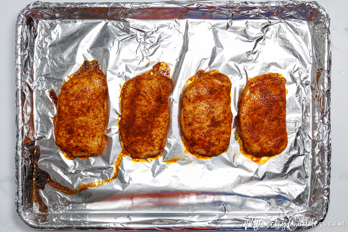 Partially baked boneless pork chops with seasoning on a foil-lined sheet pan.