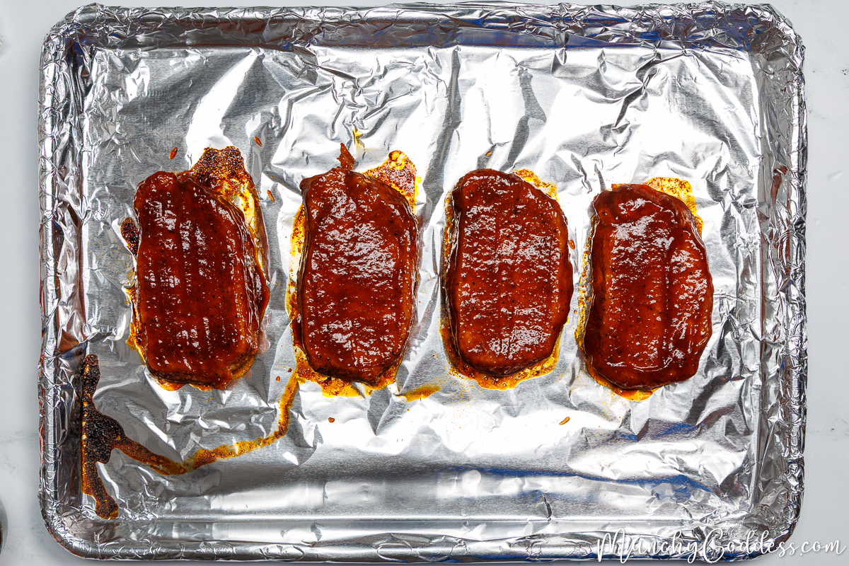 Partially baked boneless pork chops brushed with bbq sauce on all sides on a foil-lined sheet pan.