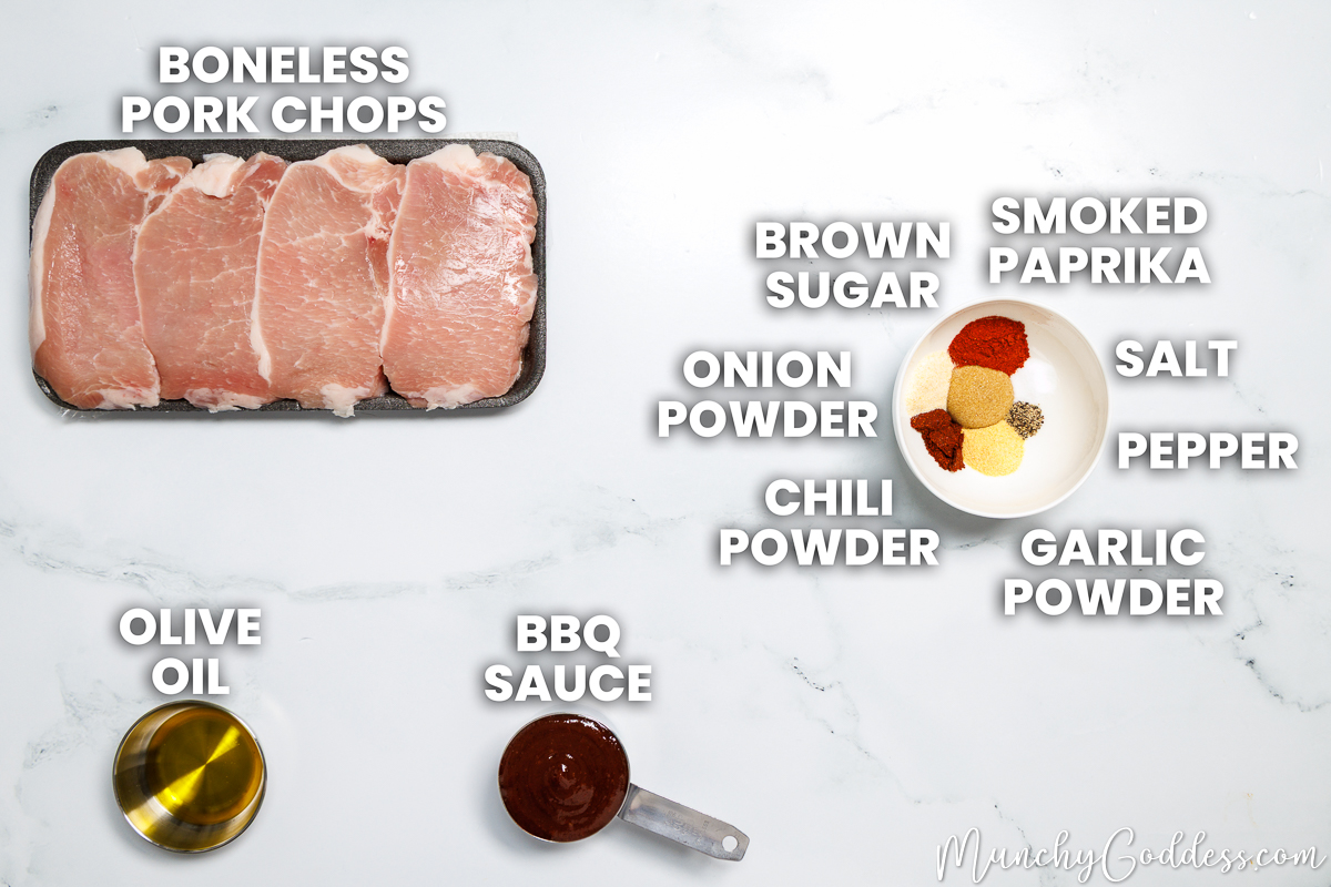 Baked bbq pork chops recipe ingredients in various dishes on a white marble surface: boneless pork chops, olive oil, bbq sauce, brown sugar, smoked paprika, salt, ground black pepper, garlic powder, chili powder, and onion powder.