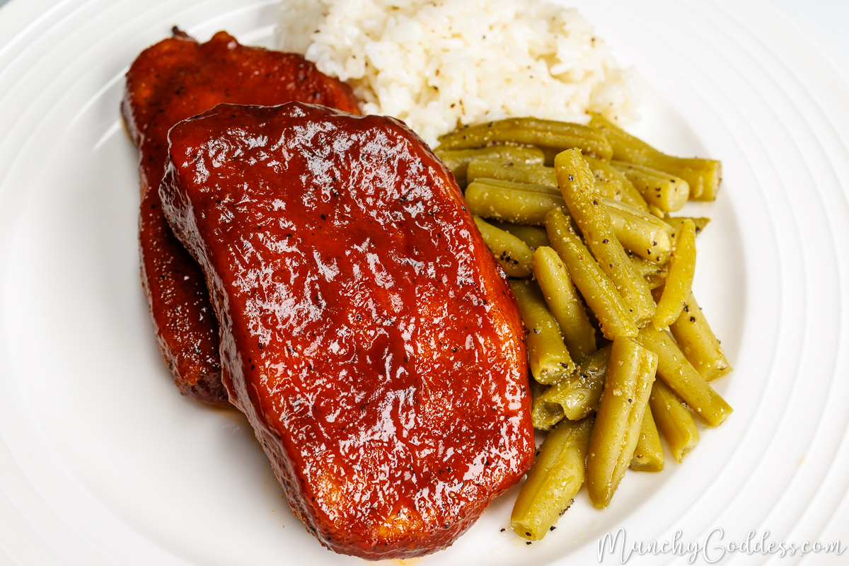 Baked bbq pork chops served with green beans and rice on a round off-white plate.
