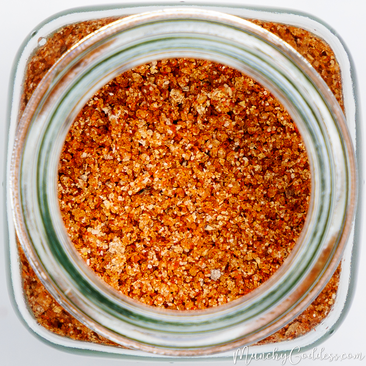 Close-up, overhead shot of a coarse BBQ spice rub inside a glass jar, featuring a mix of reddish-orange granules and visible coarse salt.