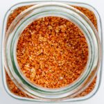 Close-up, overhead shot of a coarse BBQ spice rub inside a glass jar, featuring a mix of reddish-orange granules and visible coarse salt.
