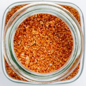Close-up, overhead shot of a coarse BBQ spice rub inside a glass jar, featuring a mix of reddish-orange granules and visible coarse salt.