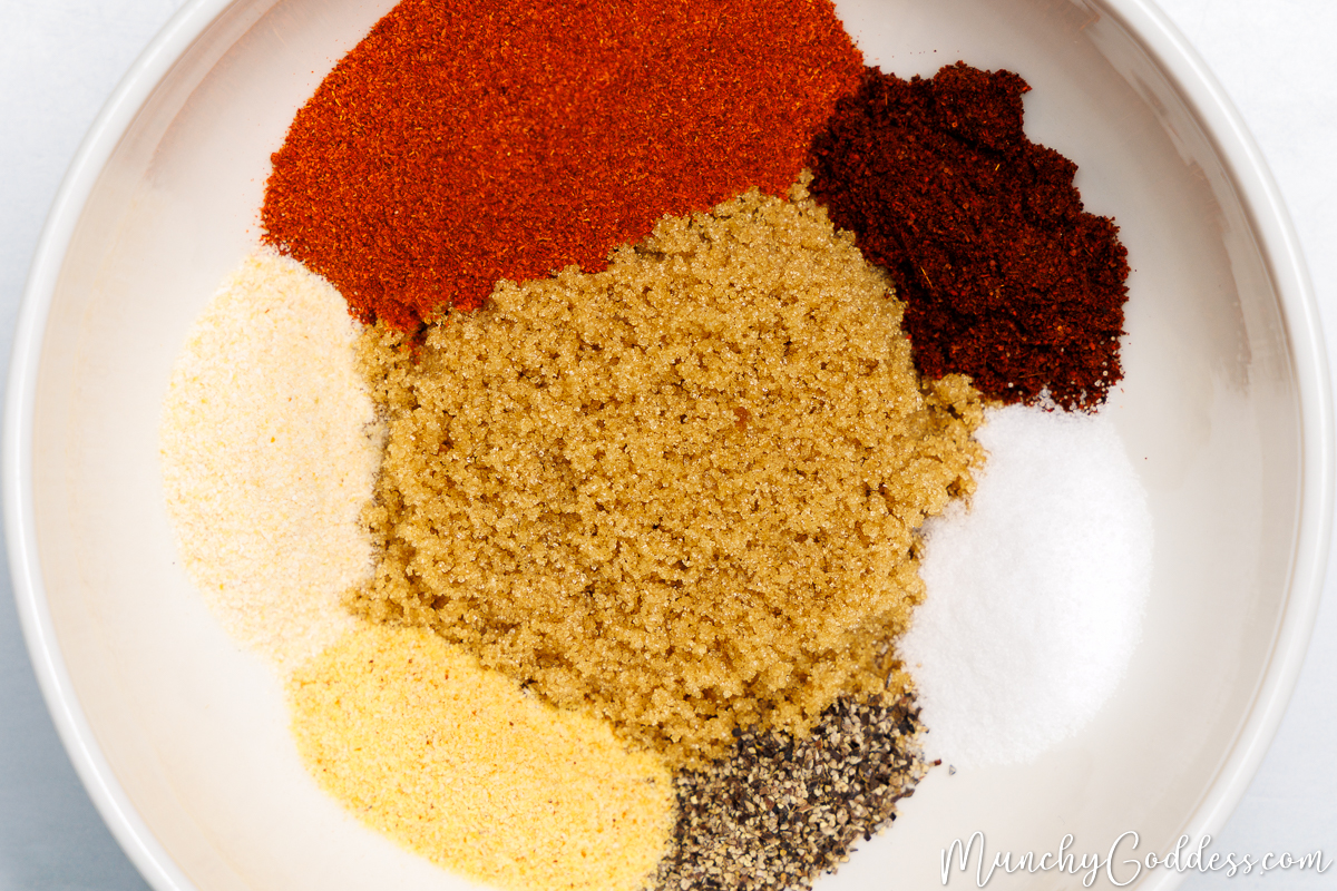 Smoked paprika, chili powder, brown sugar, salt, ground black pepper, garlic powder, and onion powder neatly arranged in an off-white bowl.