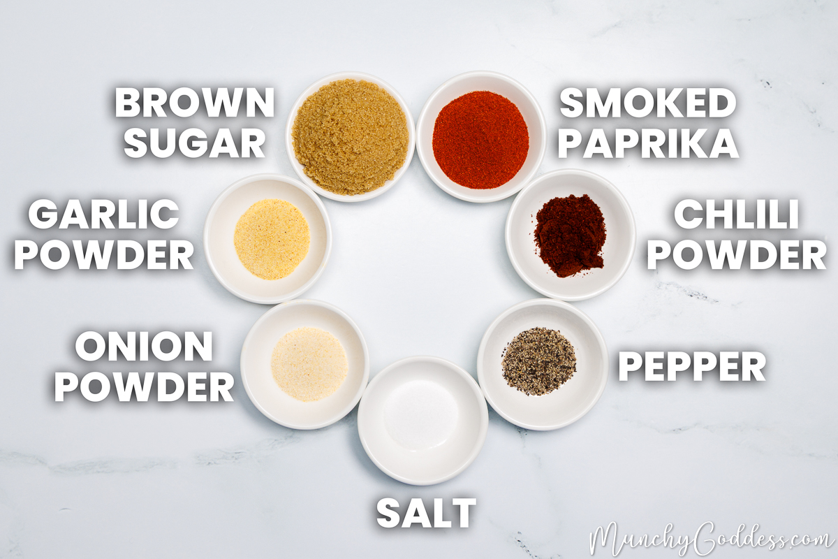 Brown Sugar, smoked paprika, chili powder, ground black pepper, salt, onion powder, and garlic powder, each in a round spice dish, with the spice dishes arranged neatly in a circle on a white marble surface.