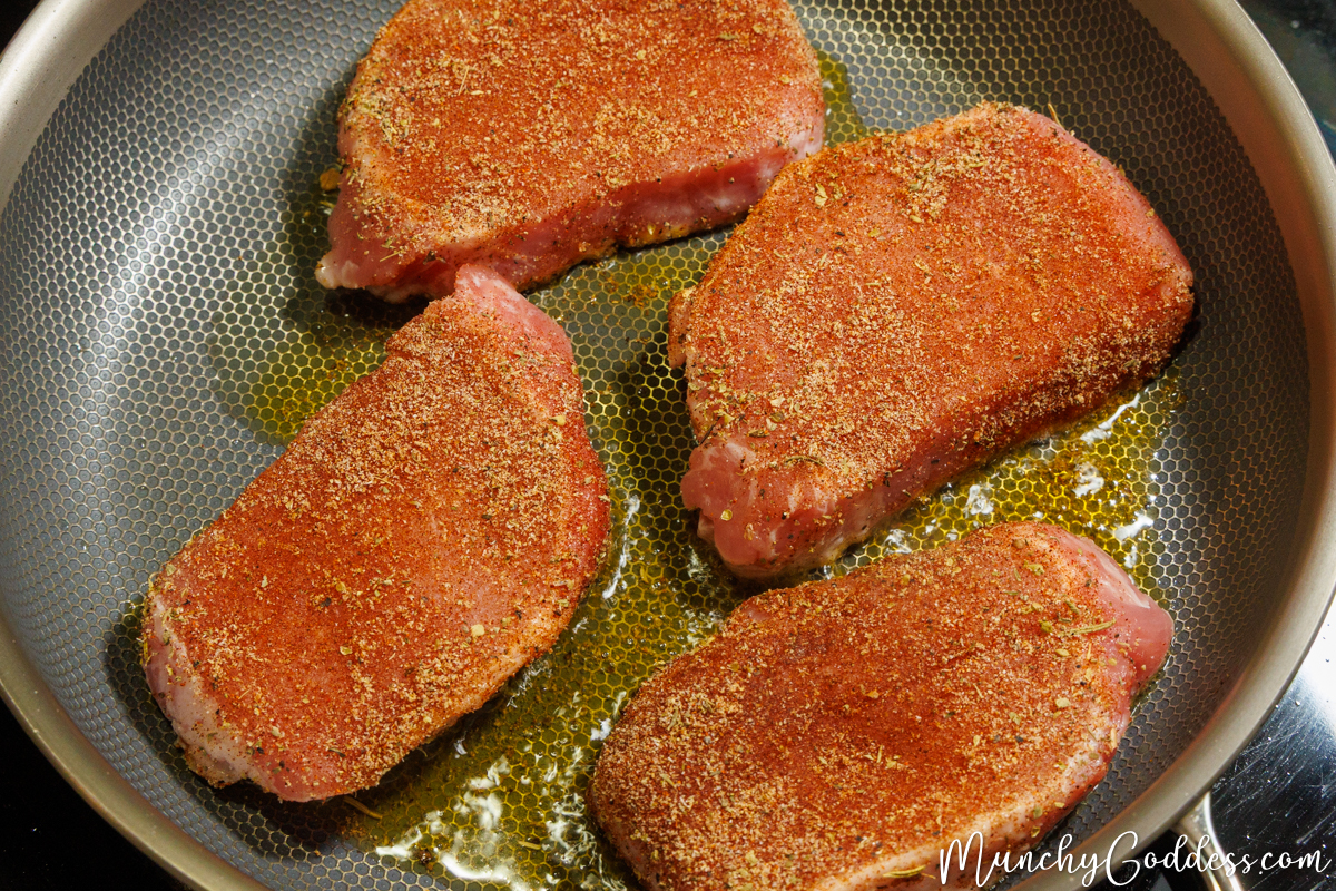 Four seasoned pork chops cooking in a skillet with olive oil.