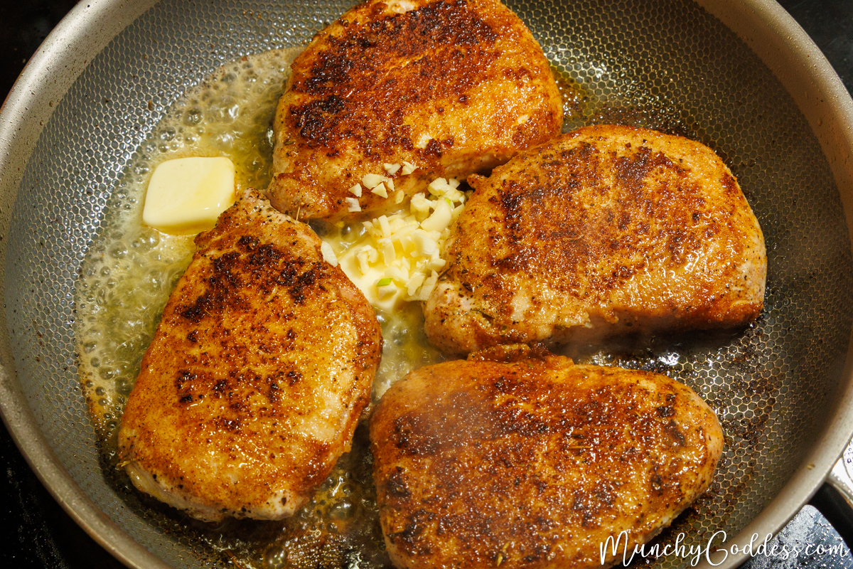 Four browned, seasoned pork chops sizzling in a skillet, with fresh minced garlic and butter melting between them.