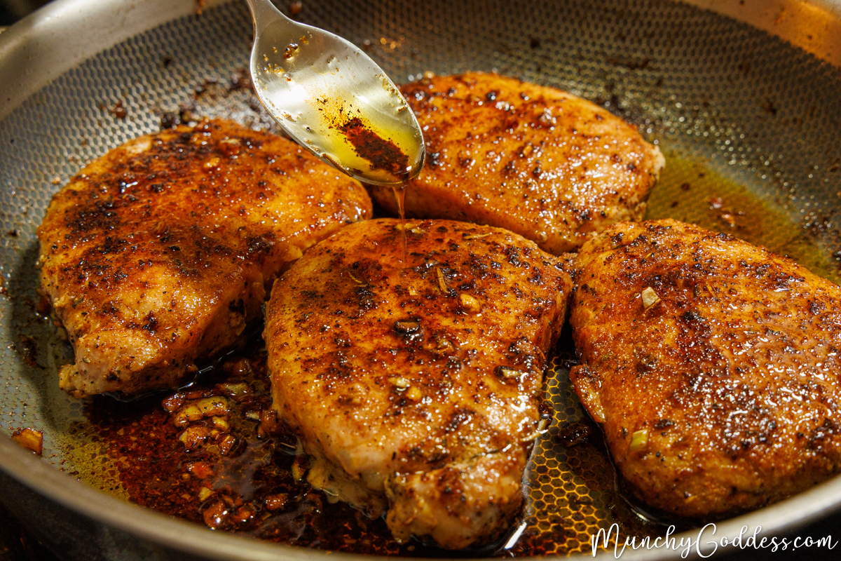 A spoon basting four finished, browned pork chops with garlic butter in a skillet.
