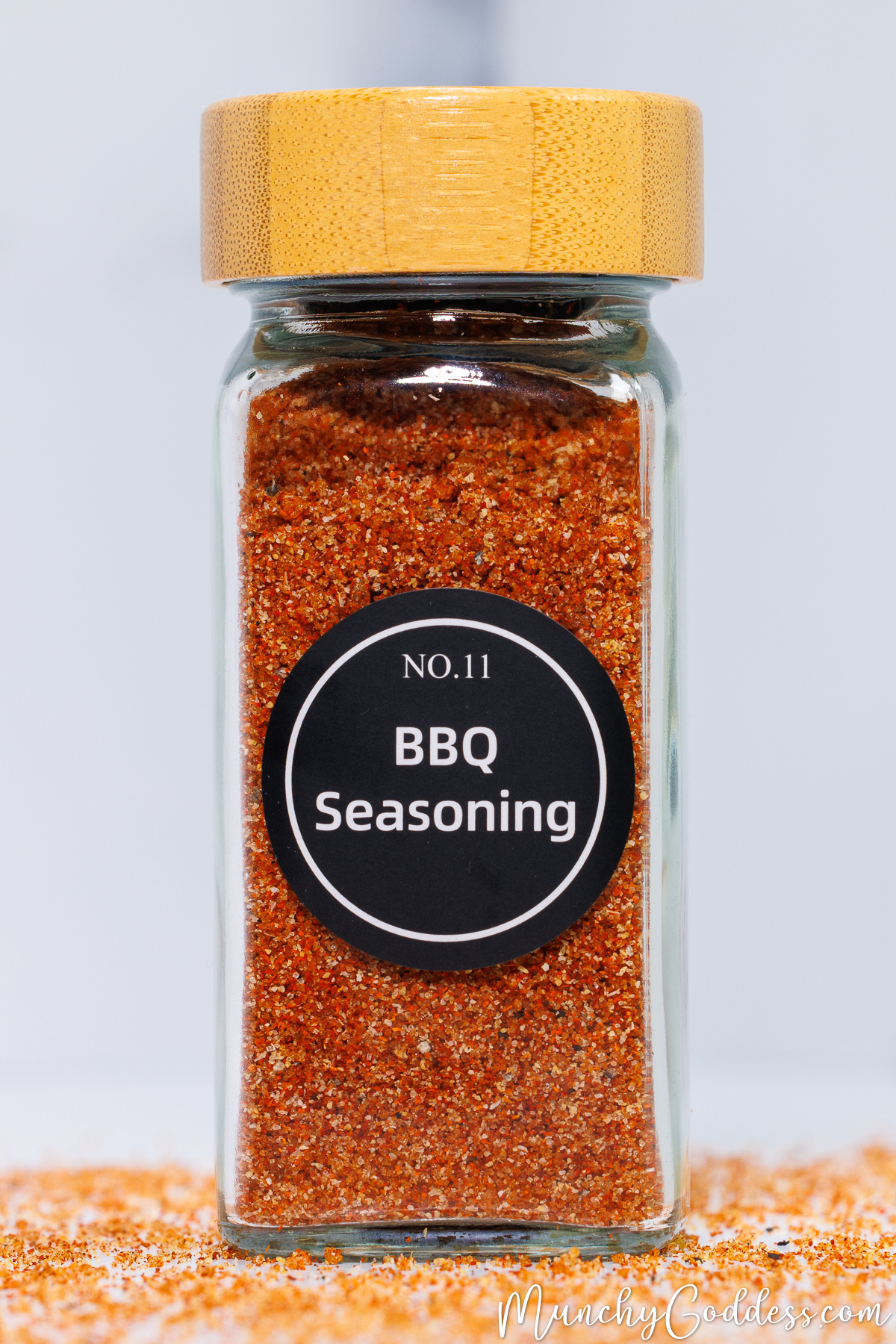 A glass spice jar with a wooden lid, filled with homemade BBQ seasoning. It features a black circular label reading 'NO. 11 BBQ Seasoning' in white text, standing on a white surface scattered with the reddish spice blend.