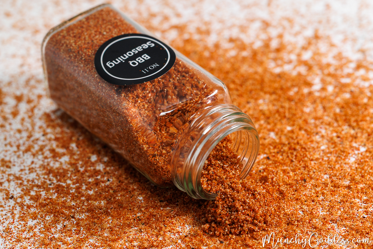Macro shot of a glass spice jar tipped on its side, spilling a vibrant, reddish-brown homemade bbq seasoning rub in a neat pile on a white marble surface.